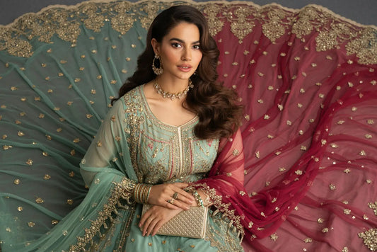 Elegant South Asian woman wearing an intricately embroidered pastel green Eid outfit with gold embellishments, styled with a contrasting red dupatta, statement jewelry, and a clutch, posing gracefully against a festive fabric backdrop.