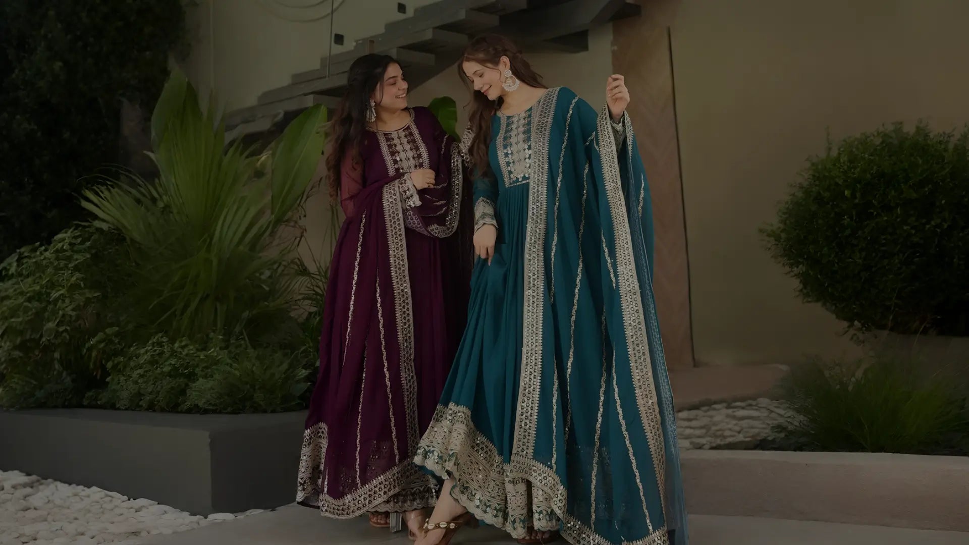 Two women in traditional outfits standing outdoors with greenery in the background