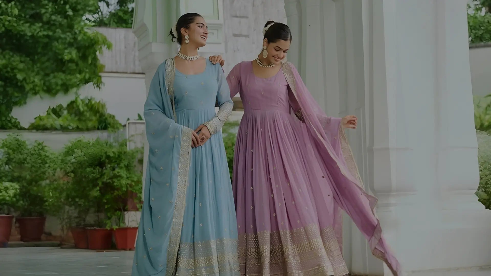 Two women in traditional attire standing outdoors with greenery and a white building in the background.