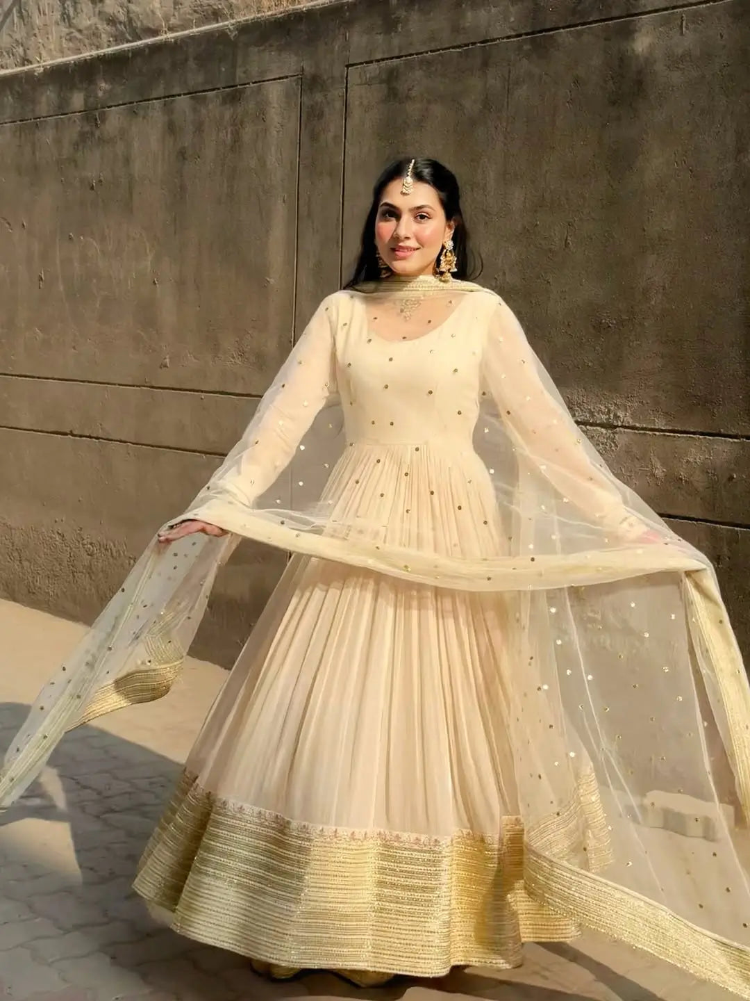 Woman in a traditional outfit with a beige and gold dress against a concrete wall.