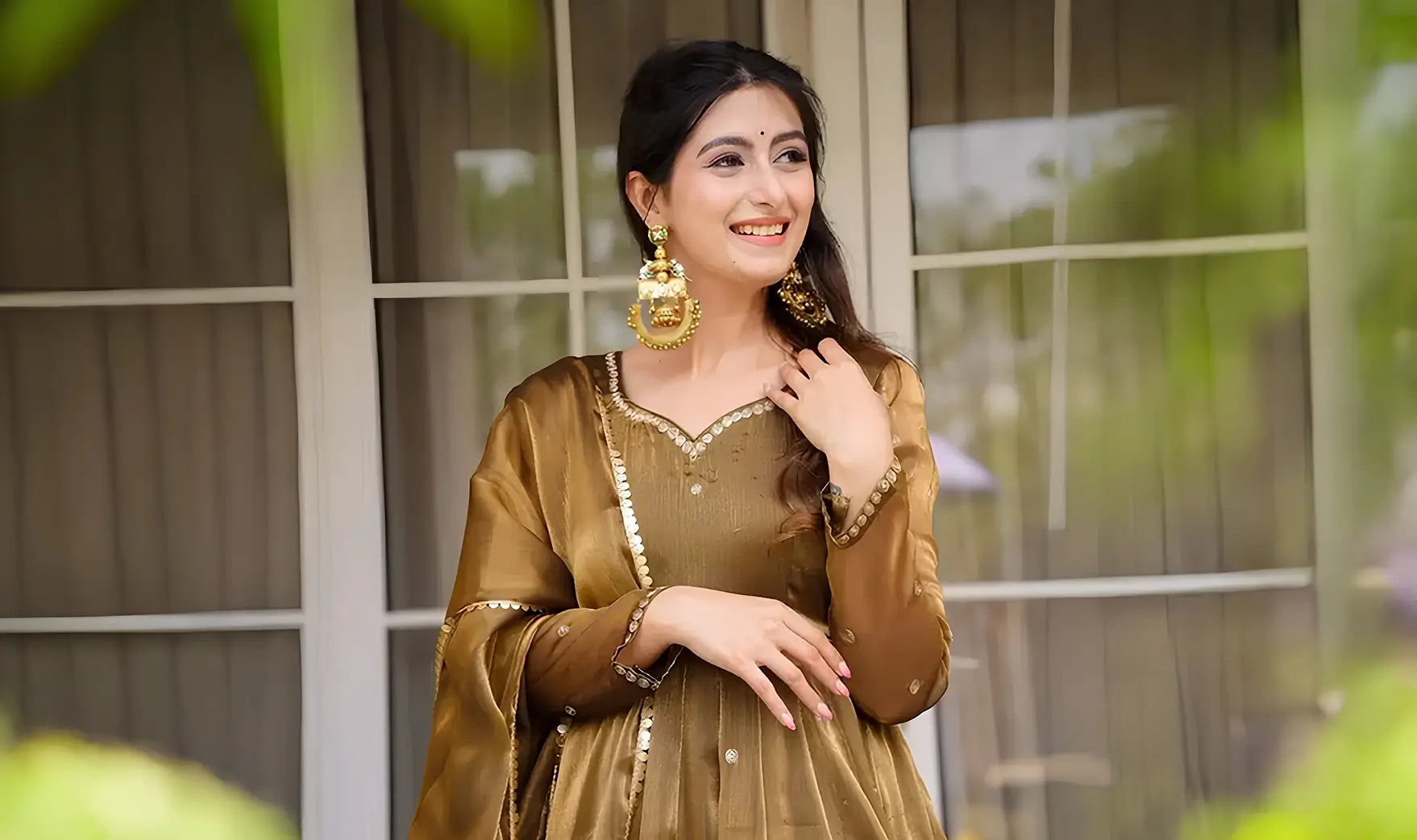 Woman in a traditional outfit with gold jewelry standing in front of a window.